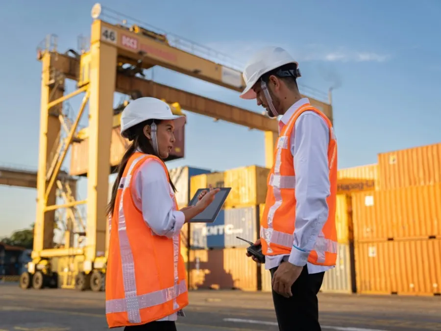 Two logistics workers in high-visibility vests checking a tablet in a container yard, with a gantry crane and stacked shipping containers behind them