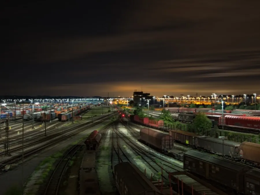 A lit freight yard at night, with stacked containers and rail wagons on parallel tracks