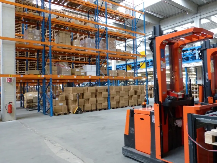 A forklift beside pallet racking in a distribution warehouse, with cartons staged on the floor ready to load