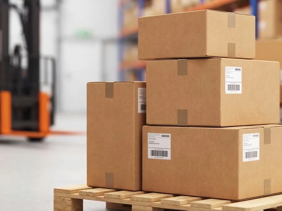 Labelled cardboard cartons stacked on a wooden pallet in a warehouse, a forklift out of focus behind them